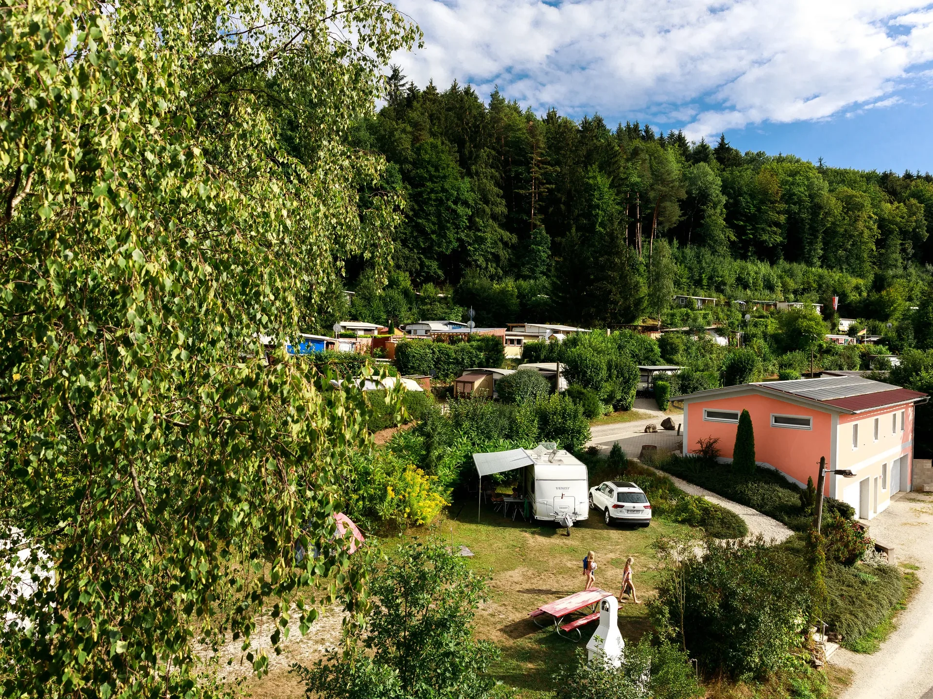 Terrassencamping mit Wohnwagen, Sanitärgebäude und bewaldetem Hang.
