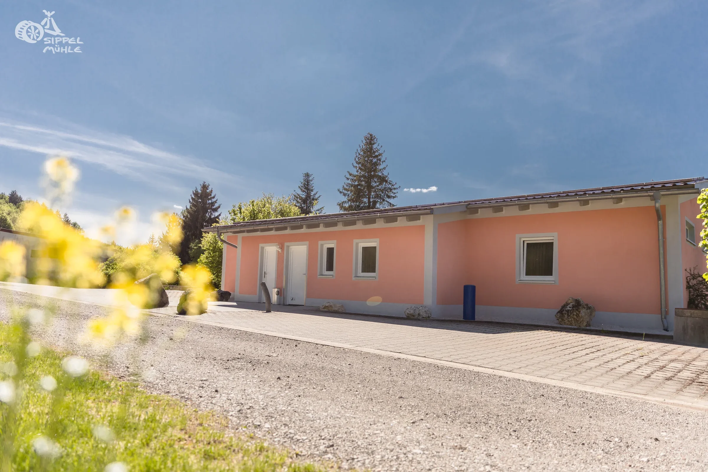 Breite Außenansicht des rosa Sanitärgebäudes mit Eingängen und gepflastertem Vorplatz.